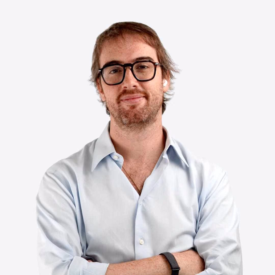 Man with beard and glasses wearing a navy blazer and white shirt in front of green foliage.