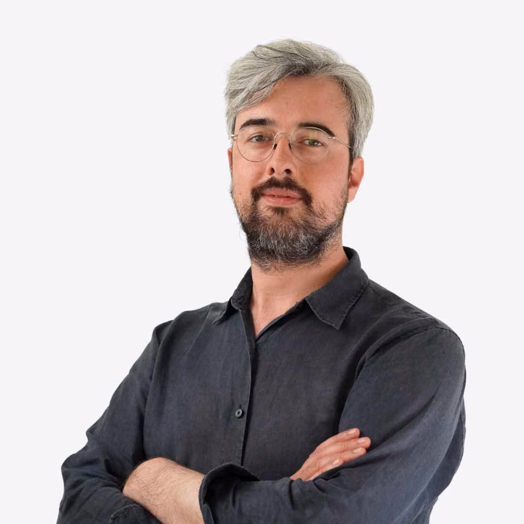 Man with gray hair and beard wearing a navy blazer and white shirt standing before green foliage.