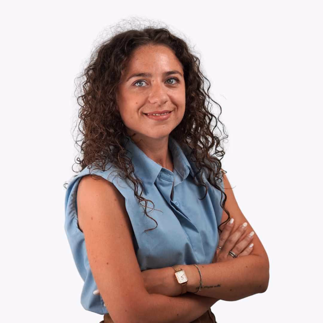 Smiling woman with curly hair wearing a blue shirt and black blazer with green foliage background.