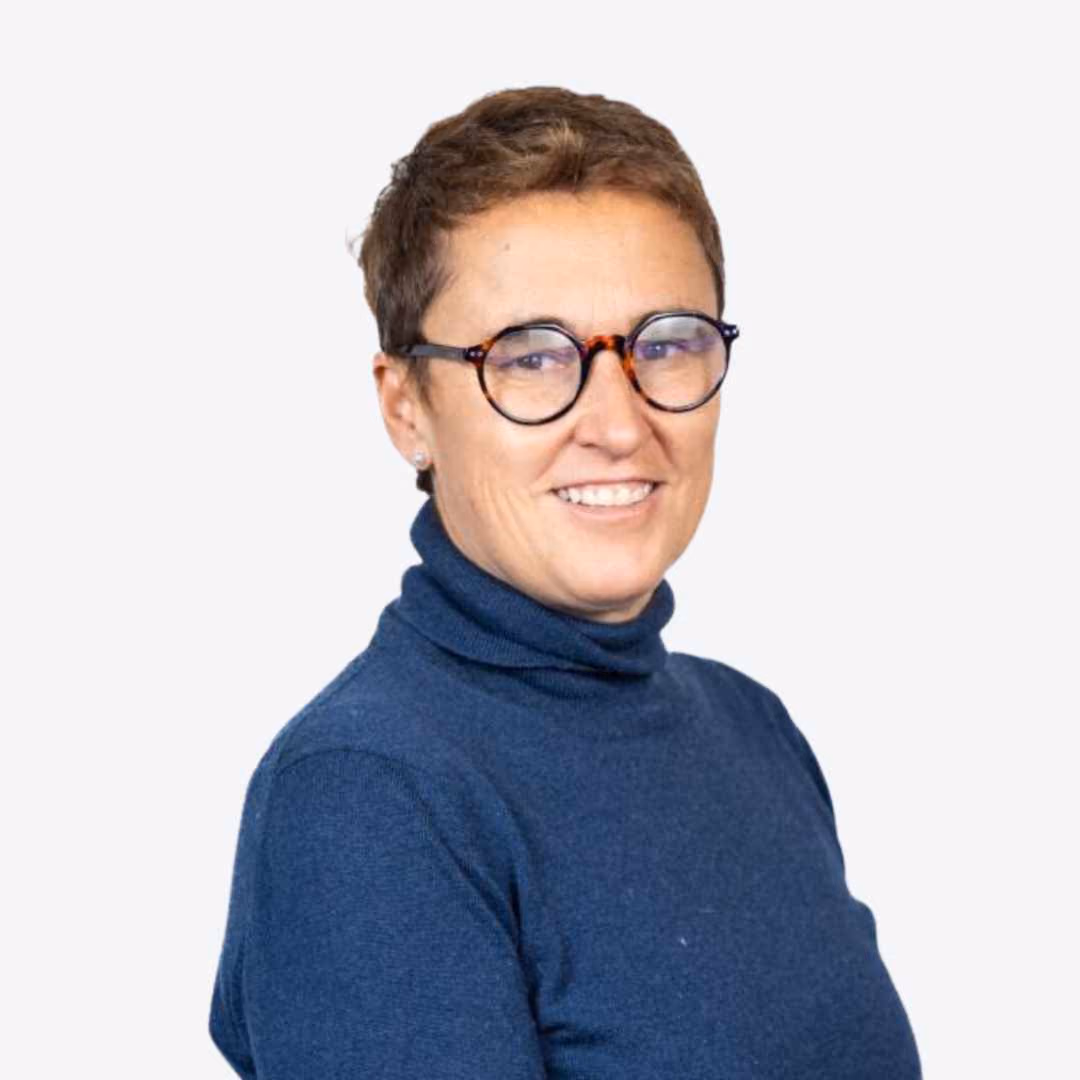 Person with short hair and glasses wearing a blue turtleneck smiling against green foliage background.