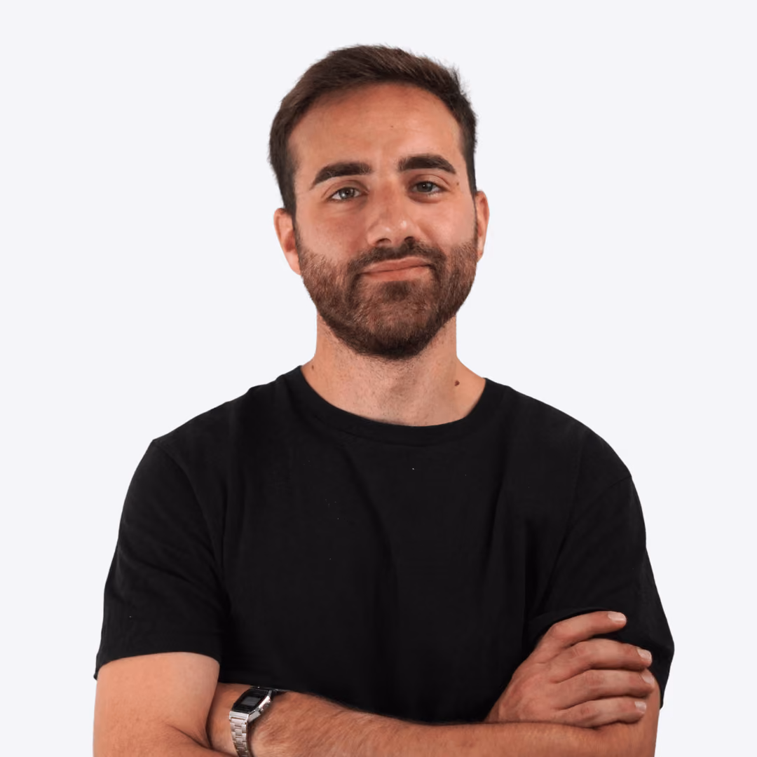 Smiling man with short dark hair and beard wearing a white shirt against a leafy green background.