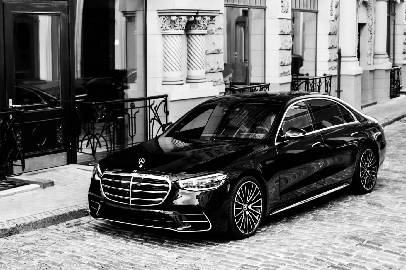 Black Mercedes-Benz S-Class parked on a cobbled street outside a stone building, black and white