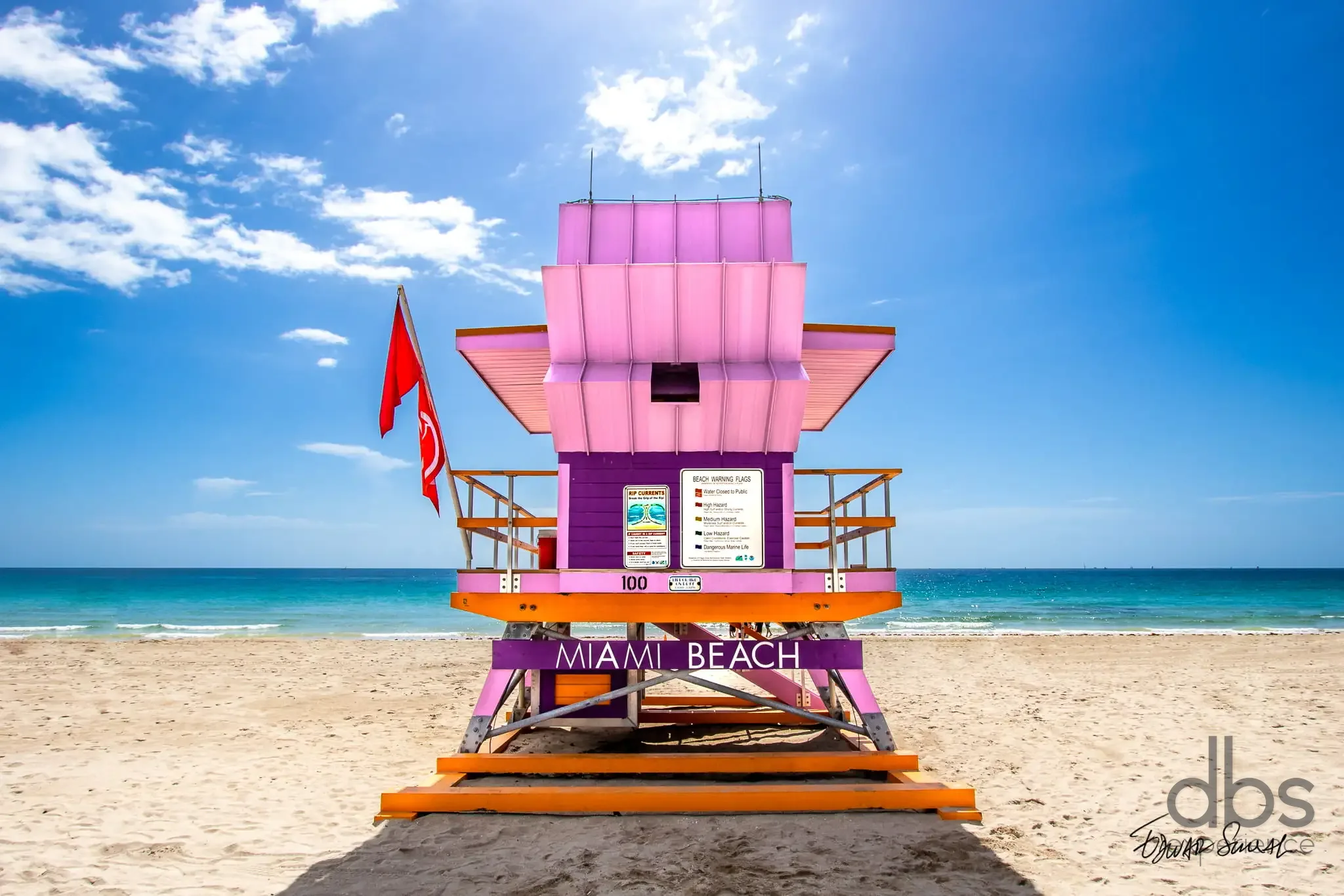 Pink and purple lifeguard tower marked Miami Beach on the sand, with a red flag and the sea behind