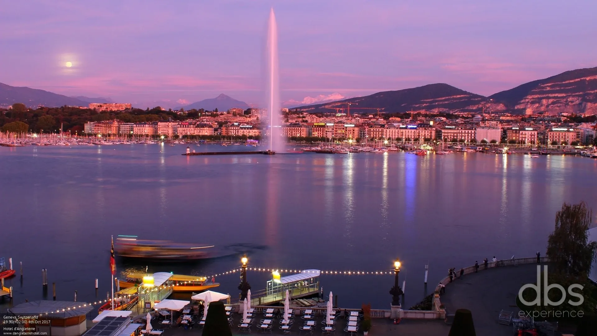 The Jet d'Eau on Lake Geneva at dusk, with the city lights and mountains behind