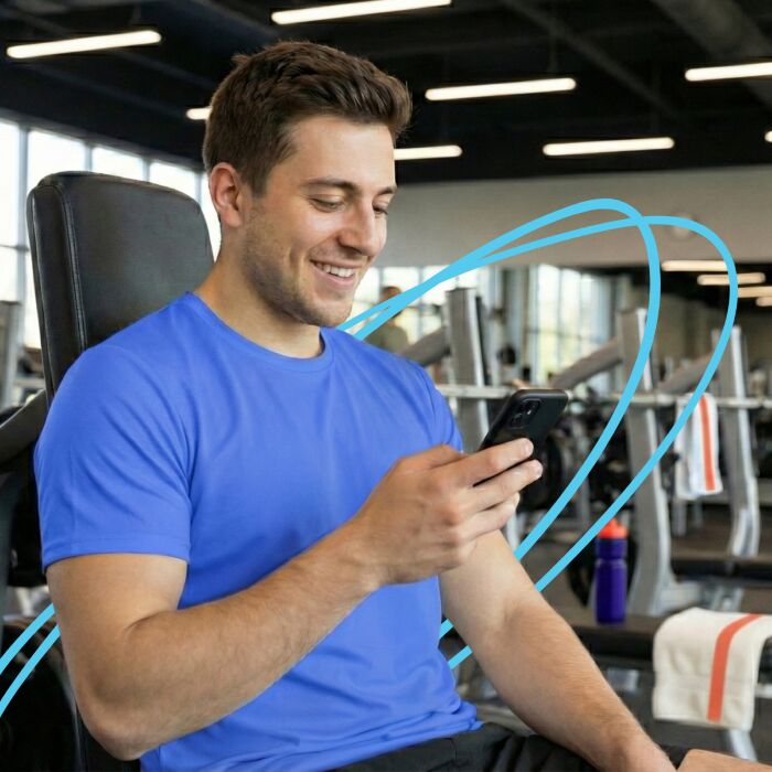 Smiling man in a blue shirt sitting on gym equipment checking his smartphone.