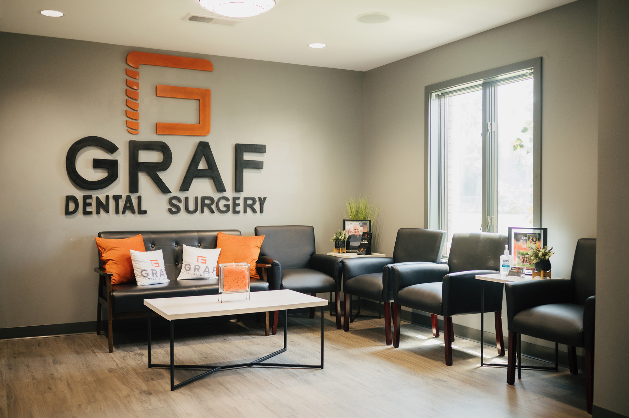 A modern dentist office waiting room with black chairs, a white coffee table, branded pillows, and "GRAF DENTAL SURGERY" signage on a gray wall.