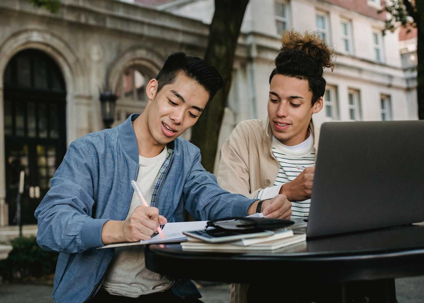 Two young men work together on a laptop at a table outside.