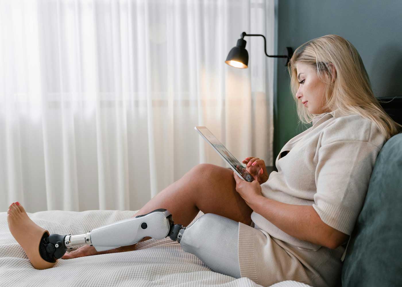 Woman with a prosthetic leg sits in bed and looks at the screen of her tablet.