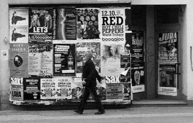 A person walks past a wall covered with various posters and advertisements, including concert and event notices, in a black and white street scene.