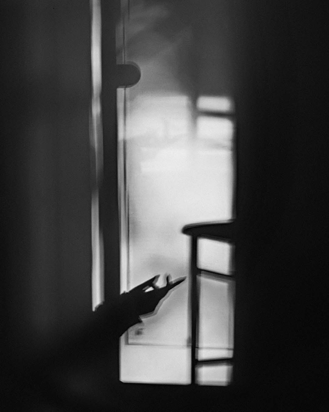 A black and white photo shows the shadow of a hand making a gesture near a window, with soft light and blurred outlines of a chair and other objects in the foreground.