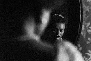 A black-and-white photo shows a woman’s face reflected in a mirror. The foreground is blurred while her face, framed by shadows, appears serious and thoughtful, standing out sharply in the reflection.