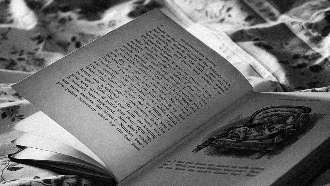 An open book rests on a patterned fabric surface. One page has printed text, while the facing page features an illustration of a person sitting in a large chair. The image is in black and white.