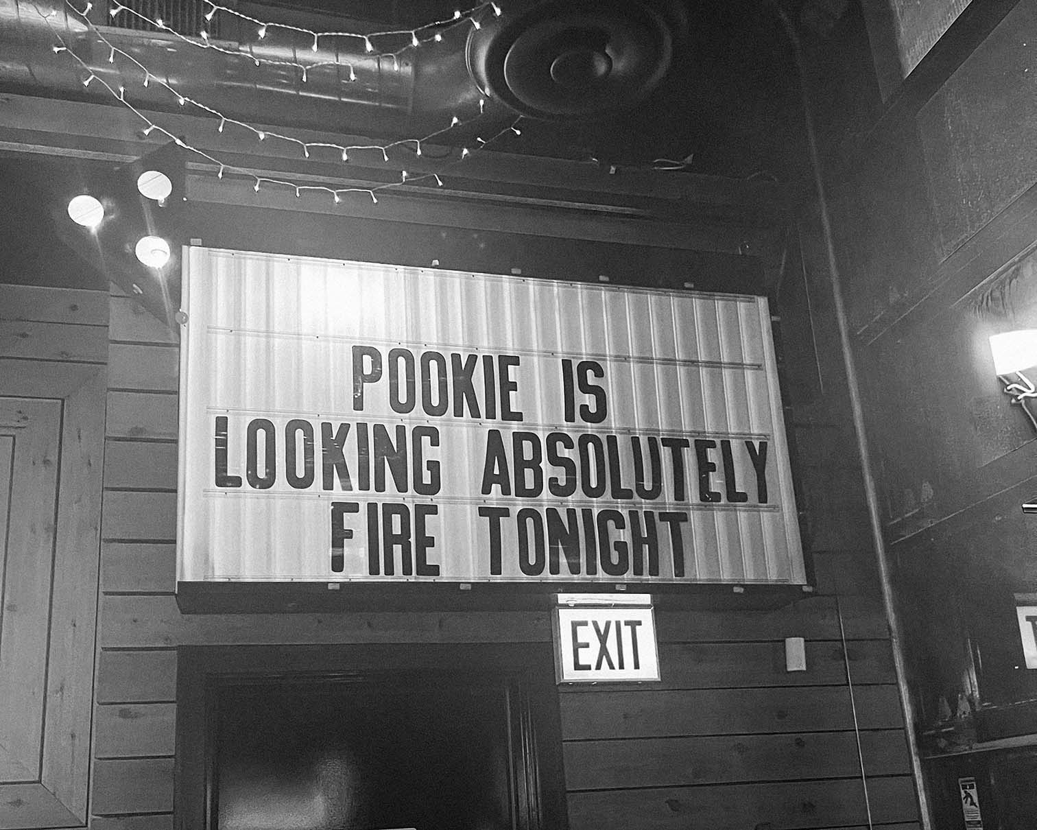 A black and white photo of a sign on a wooden wall that reads, POOKIE IS LOOKING ABSOLUTELY FIRE TONIGHT. An exit sign is visible below the sign.