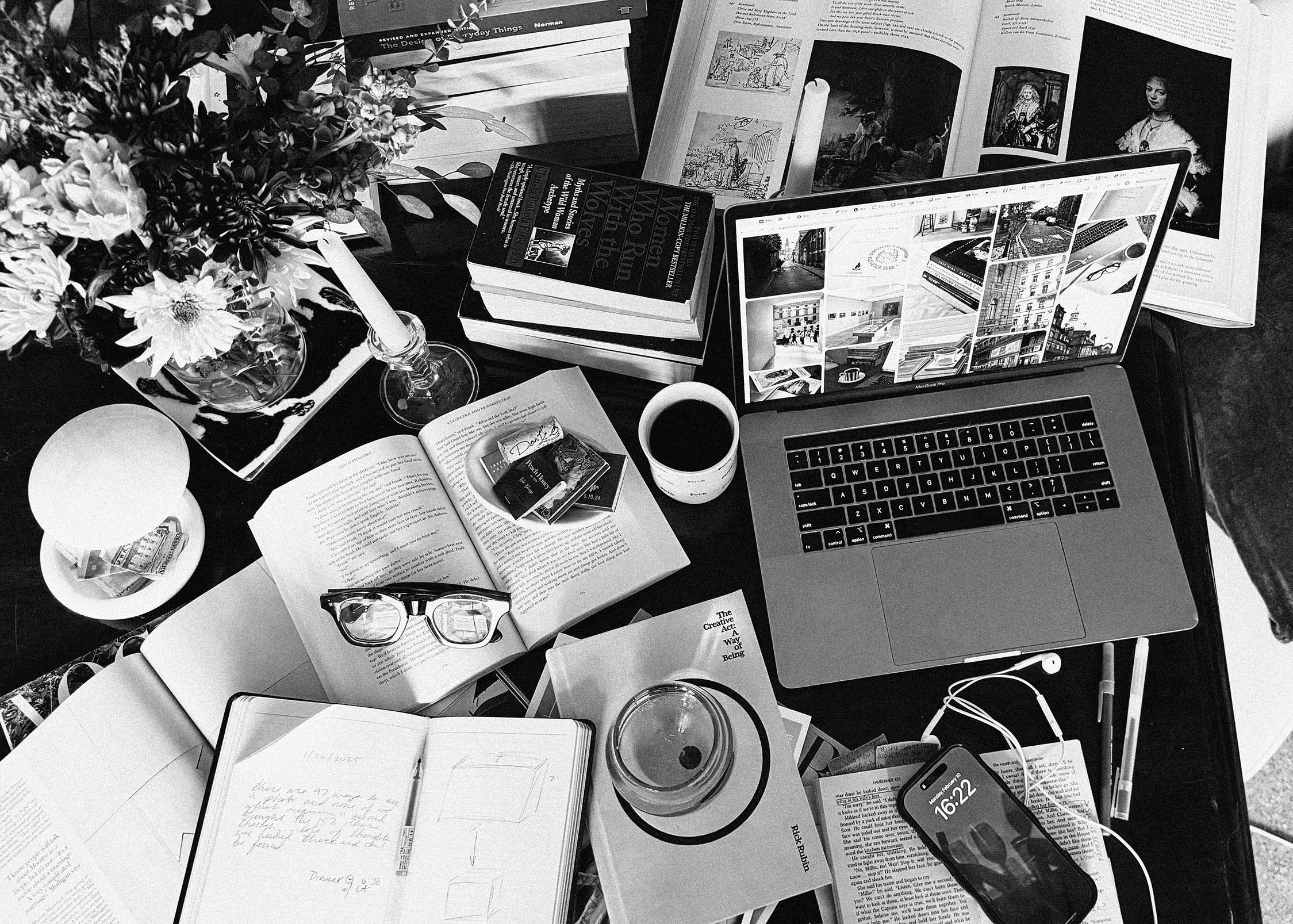 A cluttered desk with an open laptop, books, notebooks, glasses, a coffee cup, snacks, a phone, and flowers. The scene suggests intense studying or research, with various papers and items scattered around.