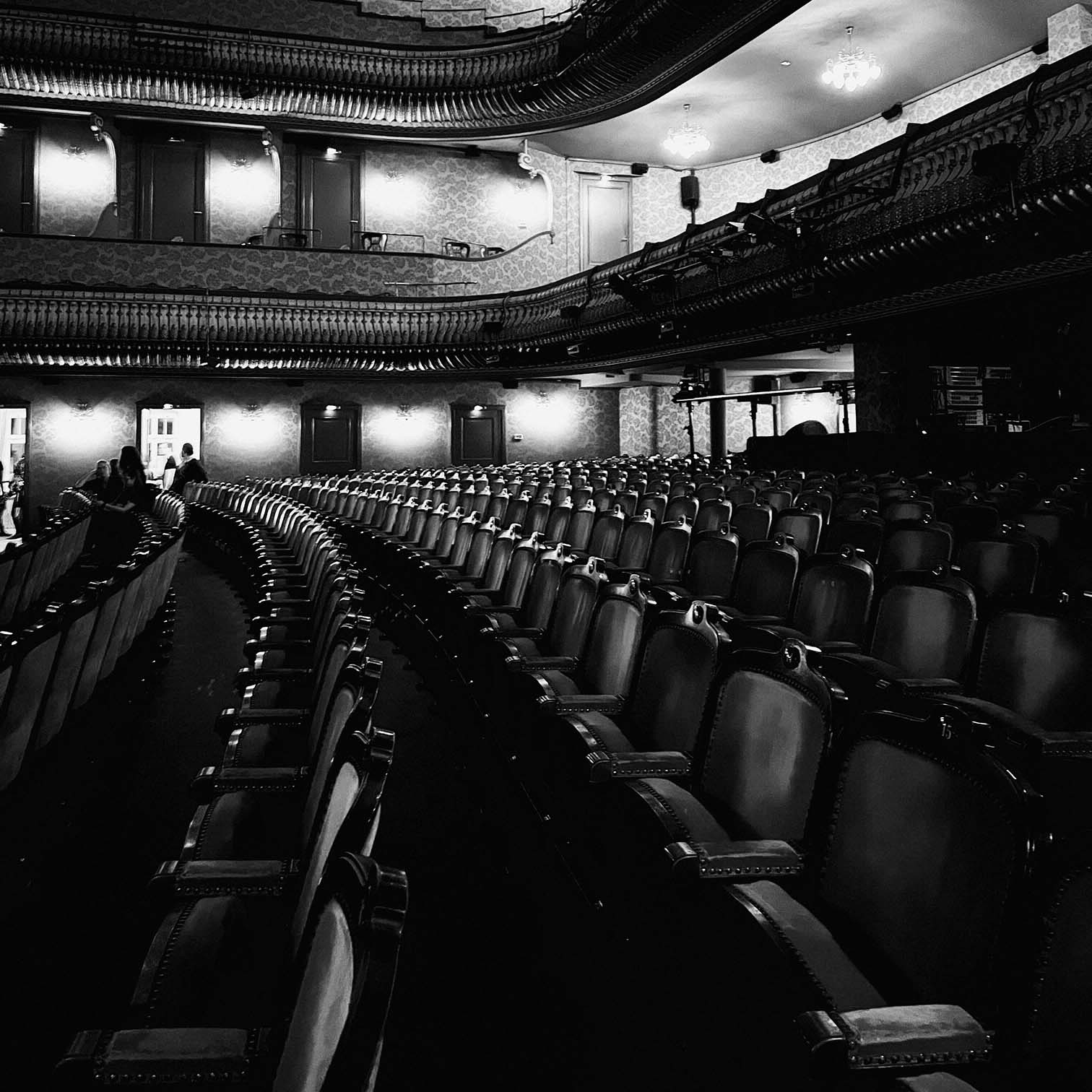 A black-and-white photo of an empty theater auditorium shows rows of ornate seats, balconies with decorative railings, and soft lighting illuminating the walls and ceiling.