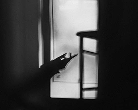 A black and white photo shows the shadow of a hand making a gesture near a window, with soft light and blurred outlines of a chair and other objects in the foreground.