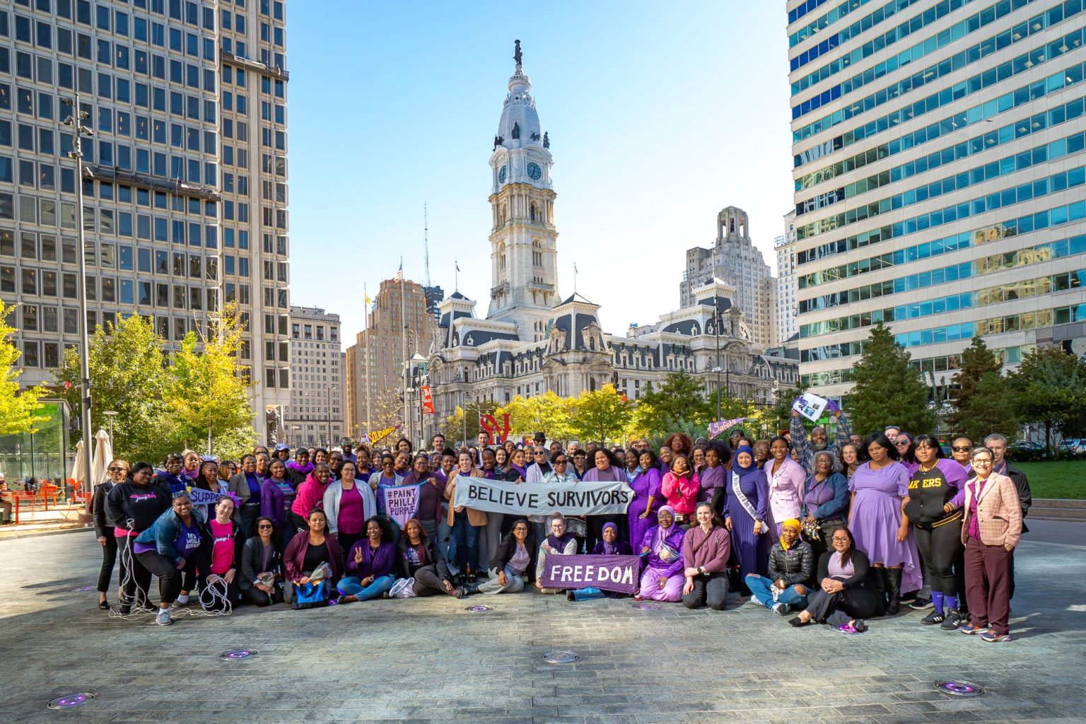 A group photo from the Paint Philly Purple event in 2025, featuring a large group of people wearing purple and smiling at the camera. City Hall is pictured in the background.