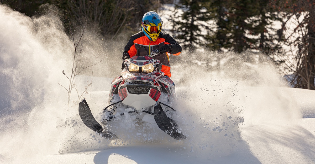 A person wearing a red and black jacket and blue helmet riding on a snowmobile with a cloud of snow behind them.