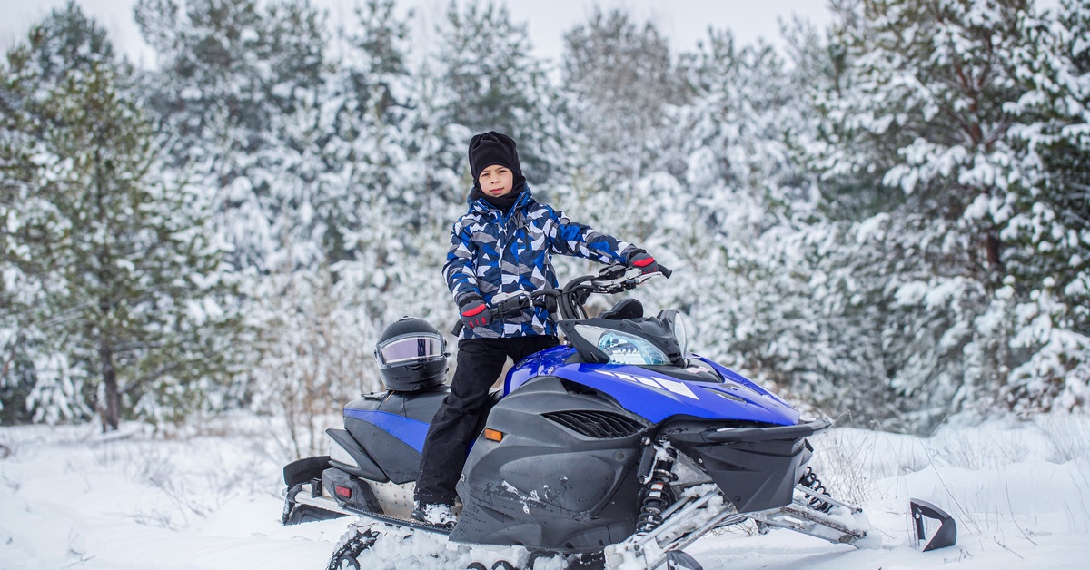 A person in a teal jacket, black snowpants, and orange helmet rides on a snowmobile alone in a vast winter landscape.