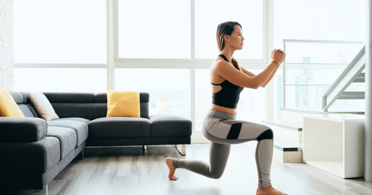 A woman in a tank top doing deep lunches in a living room with a black couch, large windows, and a wood floor.