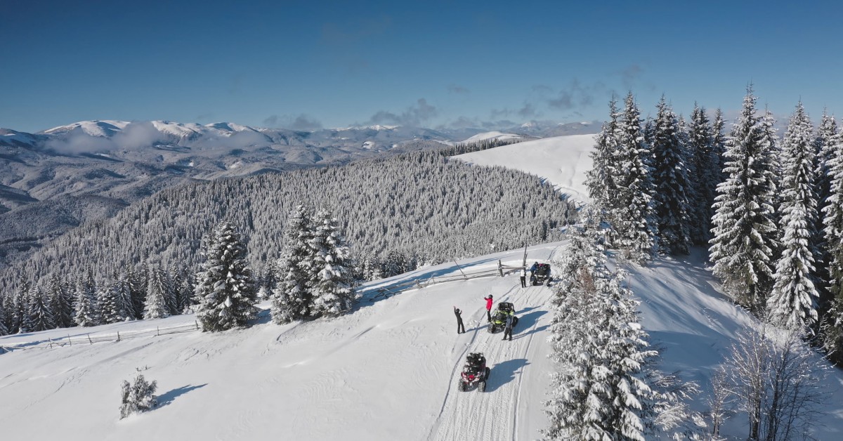 An aerial view of tourists riding snowmobiles and taking photos in a snowy mountain forest under a sunny winter sky.