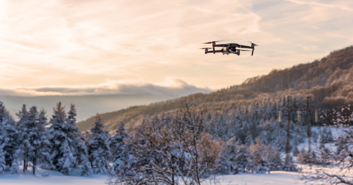 A black quadcopter drone flies over a snowy landscape with trees and hills, set against a warm-toned cloudy sky.