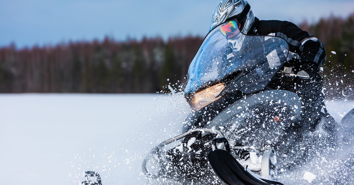 A snowmobile rider speeds through deep snow, surrounded by powder and a blurred forest in the background.