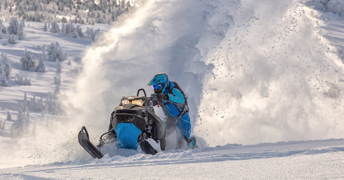 A snowmobiler makes a sharp turn, spraying snow while riding a snowmobile through a mountainous landscape.