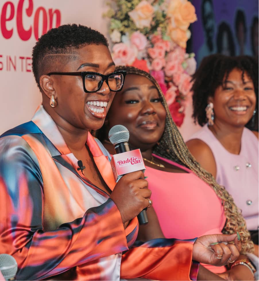 Three women smiling, one speaking into a microphone with a BaddieCon logo on it.