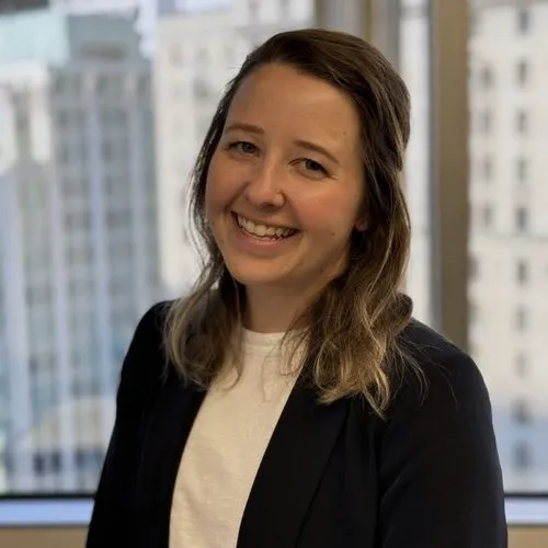Portrait of a woman smiling with light brown hair, wearing a black blazer and white top, with a blurred city background.