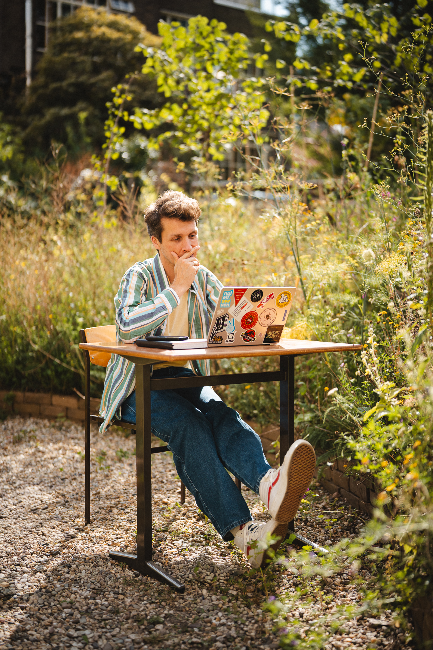 Man zittend aan bureau in tuin, verdiept in zijn laptop met stickers, omringd door groene planten.