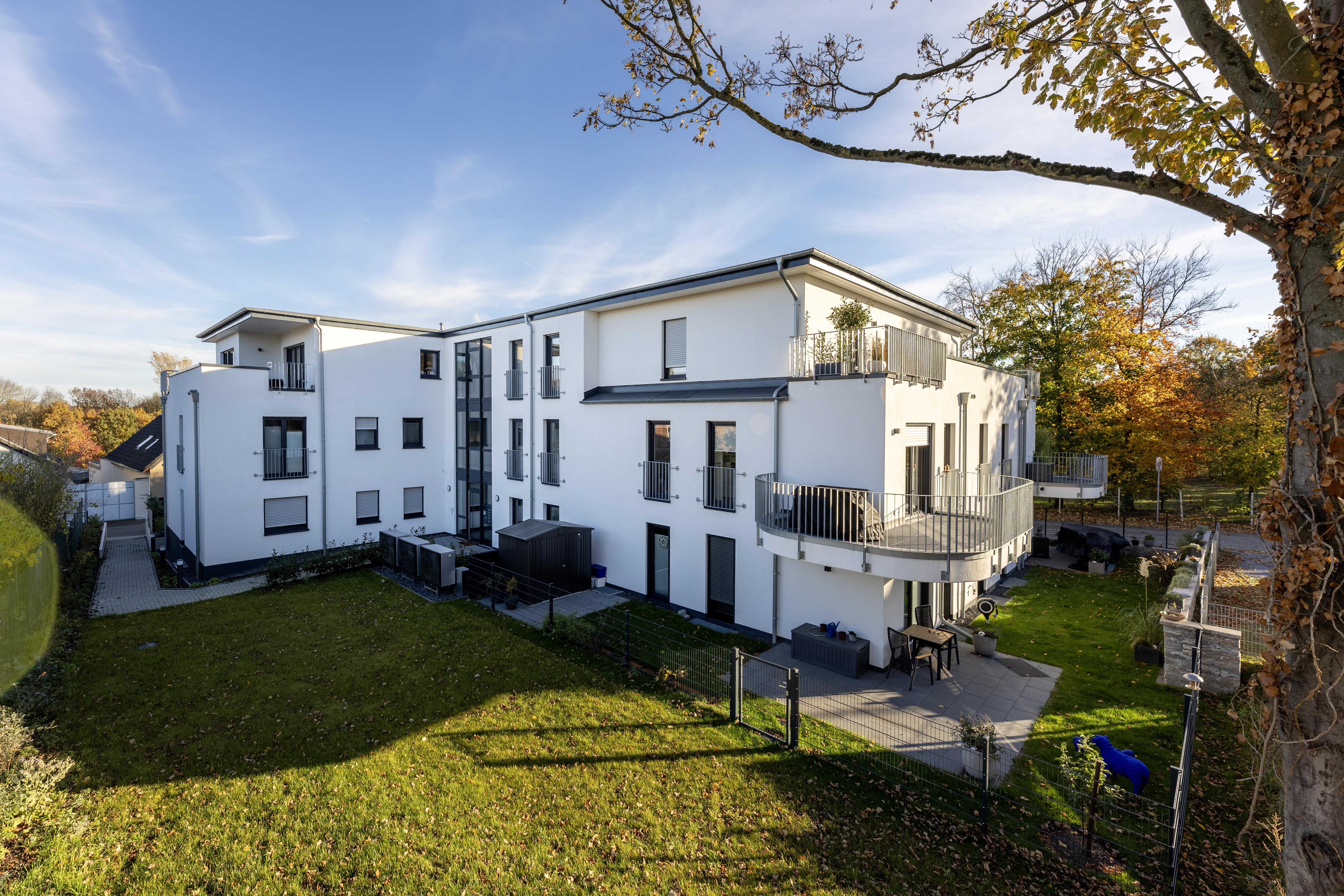 Modernes weißes dreistöckiges Wohngebäude mit Balkonen und grünem Rasen unter einem klaren blauen Himmel.