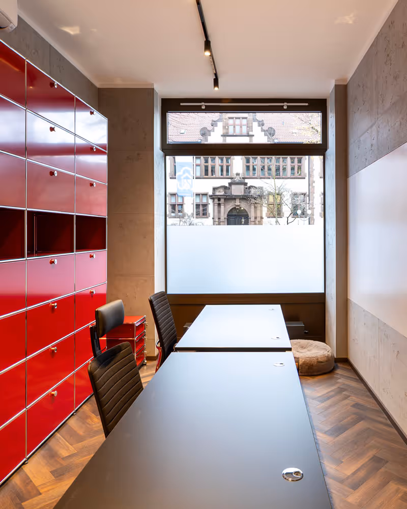 Modernes Büro mit zwei schwarzen Stühlen, langen schwarzen Tischen, roten Schranksystemen an der linken Wand und großem Fenster mit Blick auf ein historisches Gebäude.