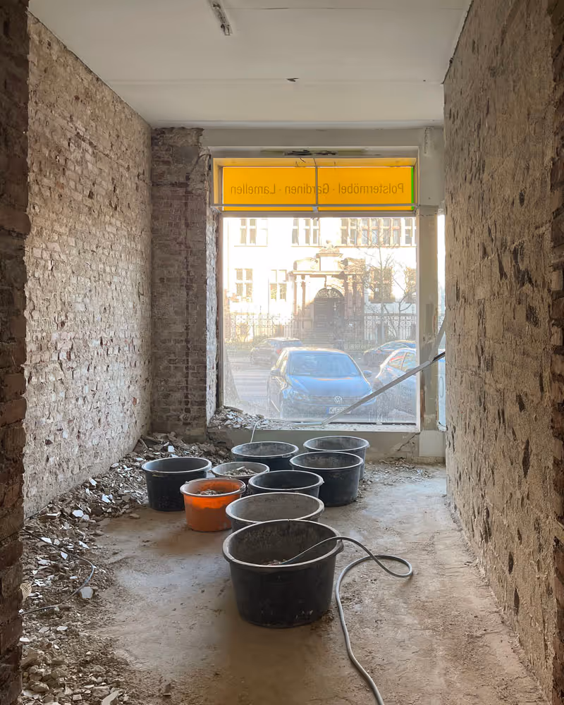 Baustellenraum mit nackten Backsteinwänden, mehreren Eimern auf dem Boden und einem großen Fenster mit gelbem Schild darüber.