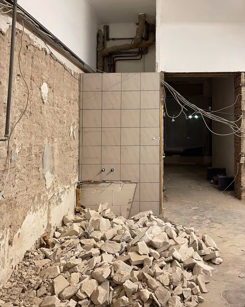 Renovierungsbereich mit freigelegter Wand, Fliesen, sichtbaren Rohren und einem Haufen Bauschutt auf dem Boden.