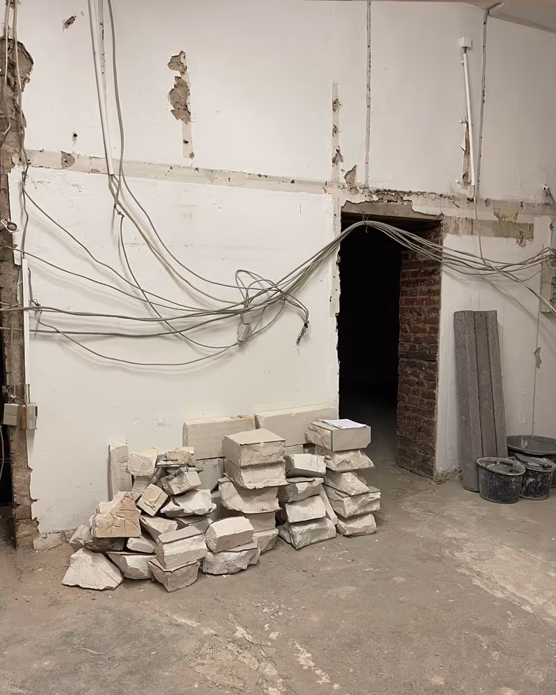 Innenraum in Renovierung mit lose gestapelten Steinblöcken auf dem Boden, freiliegenden Kabeln an der weißen Wand und einem Durchgang mit sichtbarer Ziegelwand.
