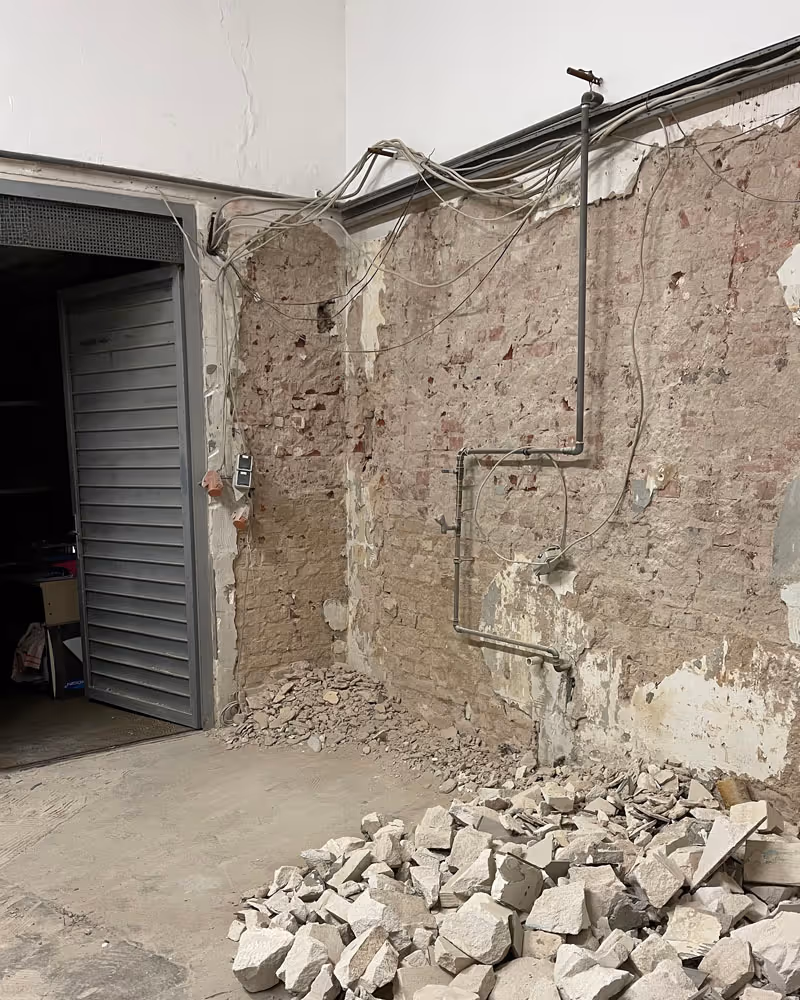 Innenraum mit abblätternder Putzwand, freiliegenden Rohren und Kabeln sowie einem Haufen zerbrochener Steine auf dem Boden.