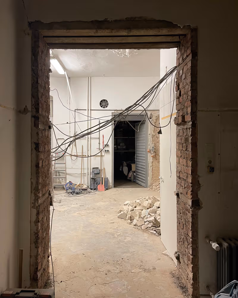 Blick durch eine roh gemauerte Türöffnung in einen renovierungsbedürftigen Raum mit freiliegenden Kabeln, Werkzeugen und einem Haufen Steine auf dem Boden.