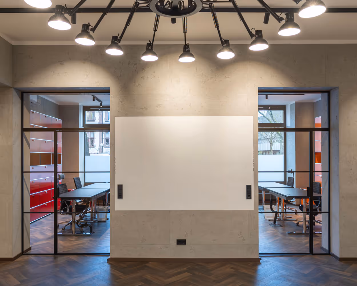 Moderner Konferenzraum mit zwei schwarzen Tischen, Bürostühlen und rotem Regal hinter Glastüren sowie weißer Tafel an der Wand.