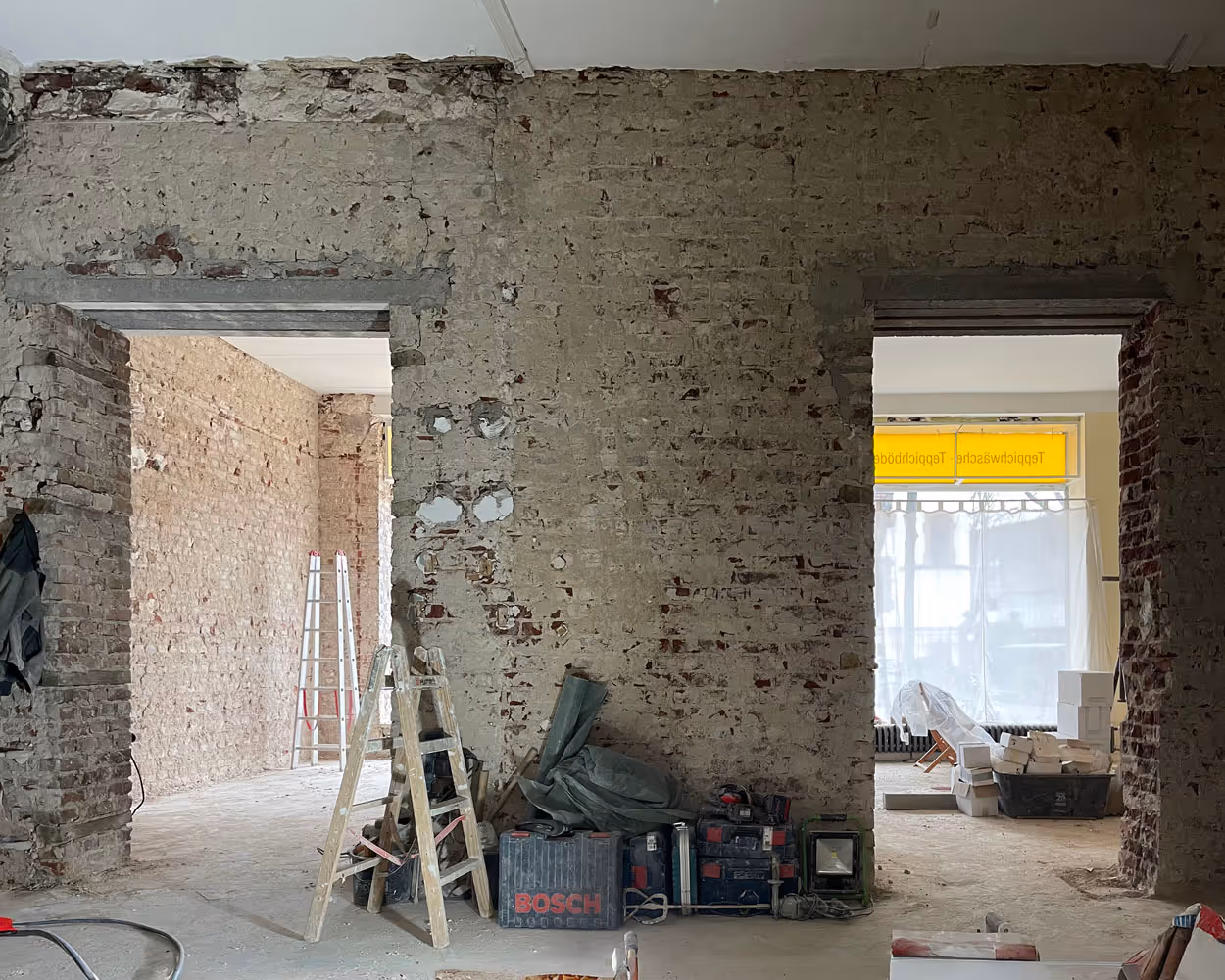 Rohbau eines Innenraums mit freigelegten Ziegelwänden, zwei offenen Türrahmen, Leitern und Werkzeugen auf dem Boden.
