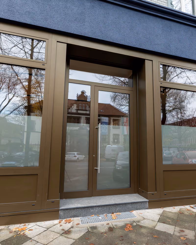 Glastür mit bräunlichem Rahmen und Bodenstufe vor einem Geschäftsgebäude mit reflektierenden Fenstern.