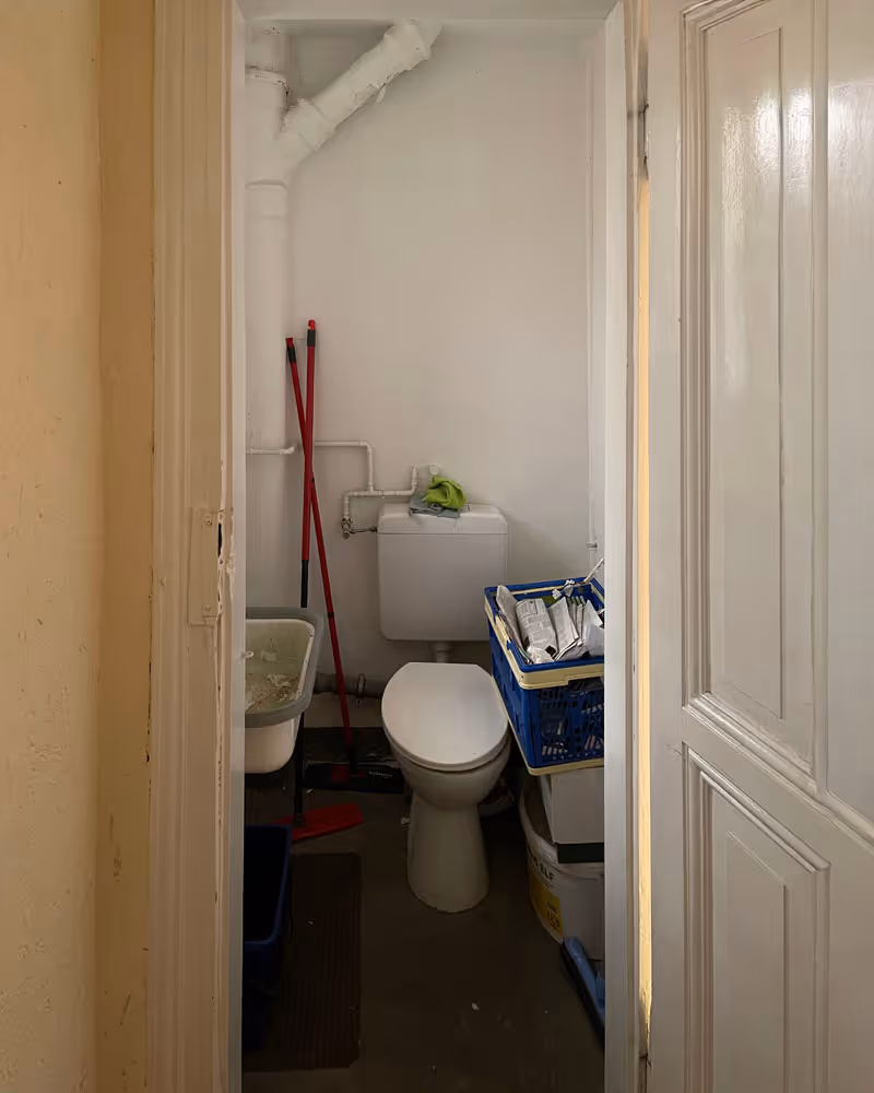 Kleines Badezimmer mit weißer Toilette, rotem Besen, Waschbecken und einem blauen Korb mit Tüten.