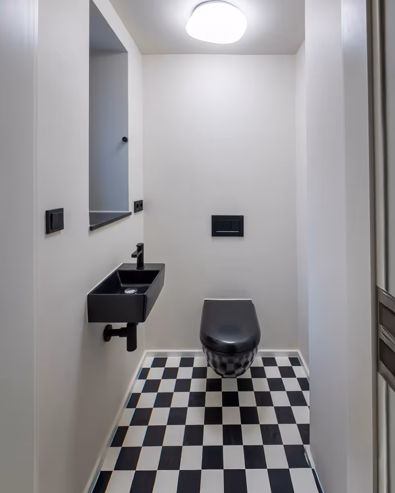 Kleines Badezimmer mit schwarz-weiß kariertem Boden, schwarzer wandhängender Toilette und schwarzem Waschbecken an weißer Wand.