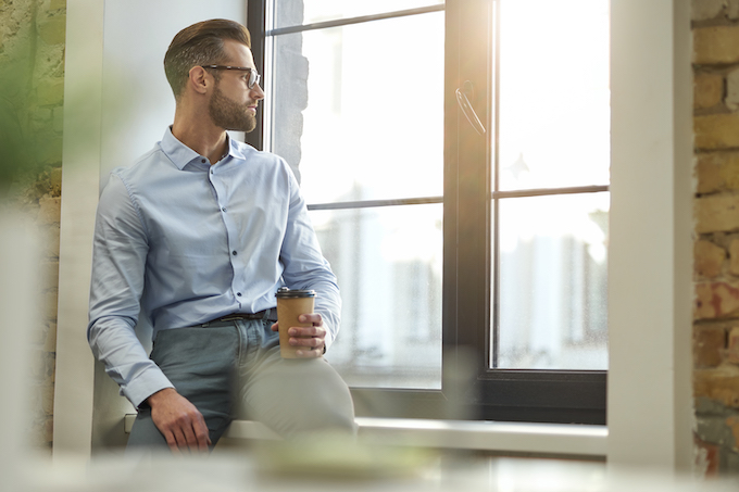 Calm young office worker sitting on a window sill with a paper cup of coffee and looking away