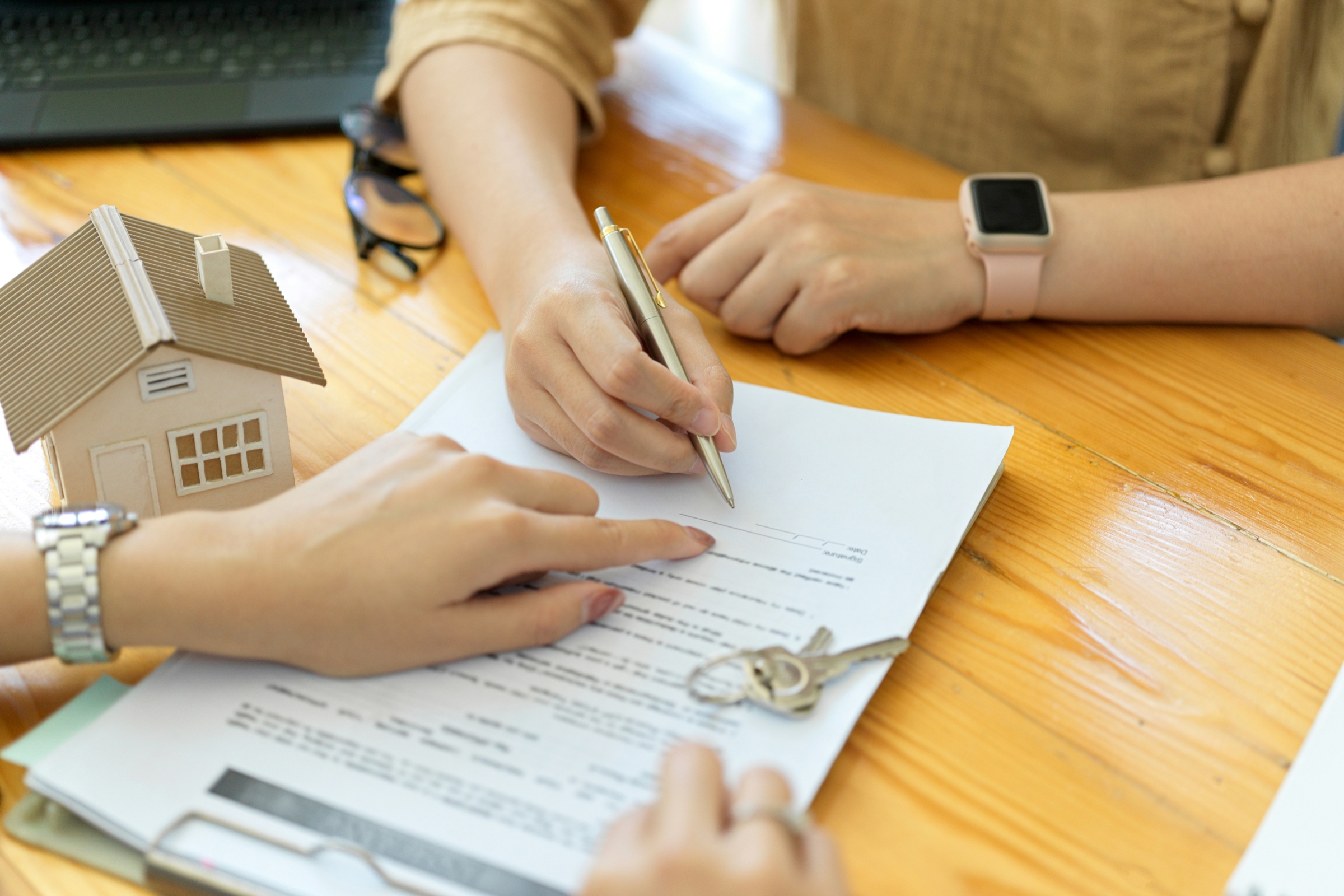 Homebuyer signing closing documents at a desk with house keys and a model home nearby