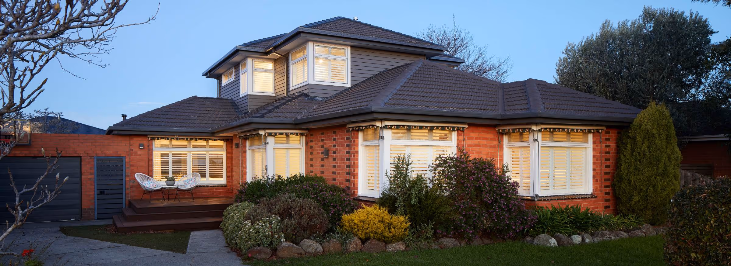A red brick home with a tiled roof and an upper-level extension, featuring white-framed windows with plantation shutters. Warm interior lights glow at dusk, and a timber deck with outdoor seating overlooks a landscaped garden.