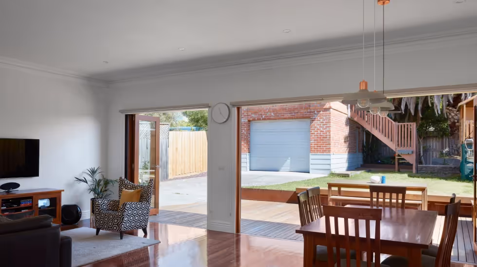 Modern open-plan living space with wooden flooring, dining area, and outdoor access through large sliding doors, leading to a backyard with a detached garage.