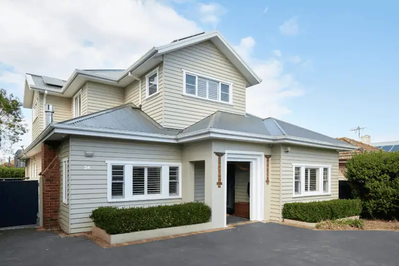 Modern two-storey weatherboard home with gable roof, brick accents, manicured hedges, and driveway under a blue sky.