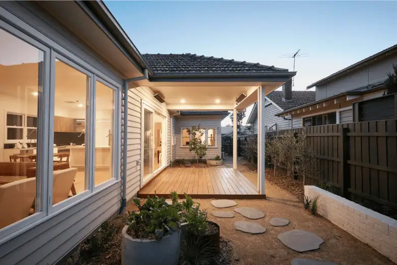Covered timber deck at the rear of a modern home, with garden beds, stepping stones, and warm interior lighting visible.