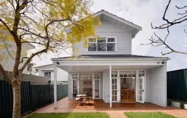Two-storey home extension with light grey weatherboard cladding, covered patio, and French doors opening to an outdoor dining area.